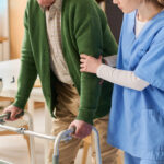 A professional home care provider using safe transfer techniques to help an elderly patient stand up from a wheelchair.